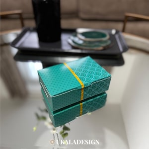 Puede incluir: Dos cajas de color azul turquesa con una franja dorada y un patrón de pequeños círculos. Las cajas están apiladas una encima de la otra.
