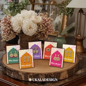 Puede incluir: Una exhibición de tarjetas de aretes coloridas sobre una losa de madera. Cada tarjeta presenta un par de aretes diferente y el texto "What Jhumka?" en verde, naranja, morado, rojo, rosa y amarillo. Las tarjetas están dispuestas frente a un jarrón de flores.