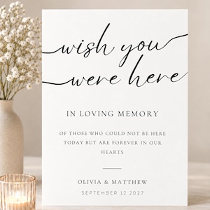May include: A white rectangular sign with black text that reads: wish you were here, IN LOVING MEMORY OF THOSE WHO COULD NOT BE HERE TODAY BUT ARE FOREVER IN OUR HEARTS, OLIVIA & MATTHEW, SEPTEMBER 12 2027. The sign features elegant script and serif fonts.