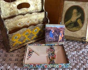 May include: A gold-painted wooden magazine rack with a decorative floral design. A small card with a religious image of Mary and Joseph riding a donkey is in front of the rack. A gold-painted picture frame with a portrait of a woman in a white dress is on the right side of the image. The items are on a white crocheted surface.