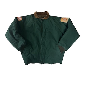 May include: A dark green bomber jacket with a brown corduroy collar. The jacket has a zipper closure and two pockets. It features a small American flag patch on the left sleeve and a patch with a fire department logo on the right sleeve.