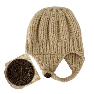 May include: A cream-colored, knitted hat with ear flaps and a button closure. The hat has a textured, ribbed design. A circular bronze emblem with an eagle design is attached to the hat.