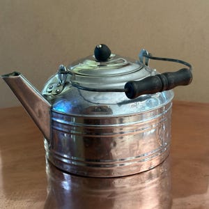 May include: A vintage metal tea kettle with a shiny silver finish. The kettle has a black wooden handle and a spout on the side. The lid is topped with a small black knob. The kettle is sitting on a copper-colored surface.