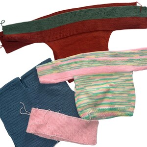 May include: A collection of unfinished knitted sweaters in various colors. The sweaters include a rust-colored one with a green neckline, a blue sweater, and a pink and pastel-colored striped sweater. A pink knitted band is also visible.