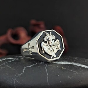 K&ouml;nnte beinhalten: Ein silberner Ring mit einem schwarzen achteckigen Gesicht, das ein detailliertes Engeldesign aufweist. Der Ring hat ein Kreuz an der Seite und ist vor einem dunklen Hintergrund platziert.