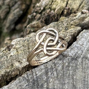 Könnte beinhalten: Ein goldfarbener Ring mit einem Knotendesign. Der Ring ist aus Metall und hat eine glatte Oberfläche.