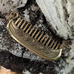 Könnte beinhalten: Ein bronzefarbener Kamm in Form eines Wikinger-Langschiffs mit detaillierten Symbolen und Designs. Der Kamm hat mehrere Zinken und runde Medaillons. Oben befindet sich eine Schlaufe zum Aufhängen. Der Kamm ist etwa 10 cm lang.