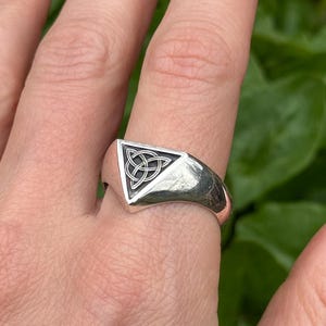 Op de afbeelding: Een zilveren ring met een driehoekig vlak met een Keltisch knoopontwerp. De ring wordt aan een vinger gedragen en toont zijn gepolijste metalen afwerking en ingewikkelde knoopwerk. De achtergrond is wazig.
