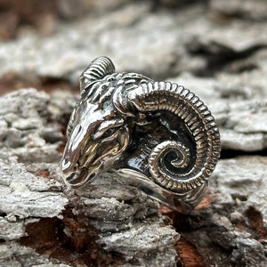 May include: A silver ring featuring a detailed ram head with curled horns. The ring is set against a textured, wooden background.