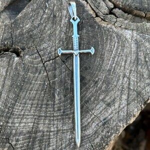May include: A silver sword pendant with a cross-shaped hilt and a pointed tip. The pendant is on a wooden surface.