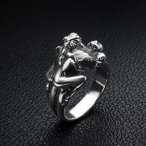 May include: A silver-colored ring featuring a sculpted design of two figures embracing. The ring has a detailed, three-dimensional appearance, with the figures' forms clearly defined. The ring is set against a dark background.