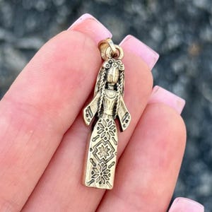 Può includere: Un ciondolo color oro a forma di donna con un lungo vestito e un copricapo. L'abito presenta un motivo floreale. Il ciondolo è tenuto da una mano con smalto rosa.