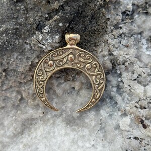 May include: A gold-colored crescent moon pendant with intricate scrollwork and dot details. The pendant has a small loop at the top for a chain and is displayed against a textured, light-colored background.