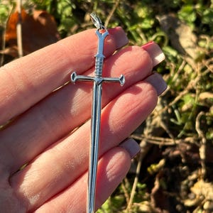 May include: A silver sword pendant with a detailed design. The pendant is about 2 inches long and has a cross-shaped guard.