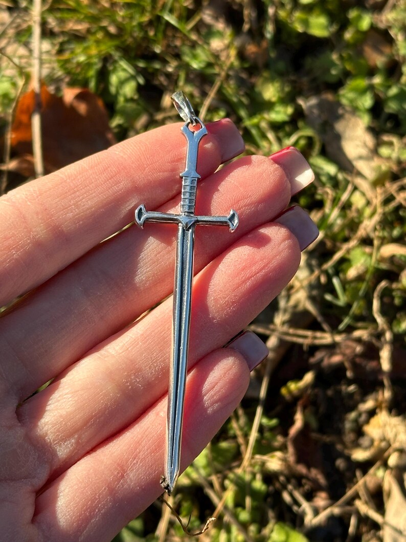 May include: A silver sword pendant with a detailed design. The pendant is about 2 inches long and has a cross-shaped guard.