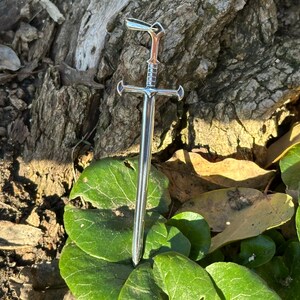 May include: A silver sword pendant with a detailed hilt and a pointed tip. The pendant is hanging from a chain and is resting on a bed of green leaves.
