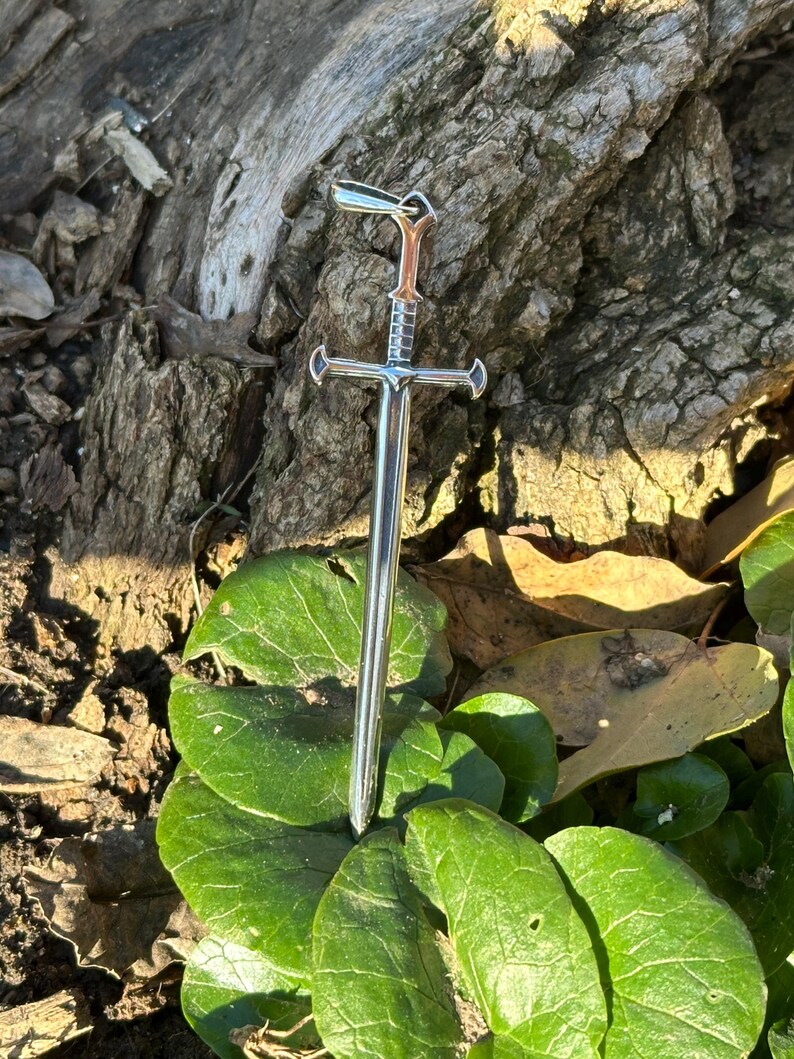 May include: A silver sword pendant with a detailed hilt and a pointed tip. The pendant is hanging from a chain and is resting on a bed of green leaves.