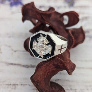 K&ouml;nnte beinhalten: Ein silberner Ring mit einer schwarzen achteckigen Fassung, die ein detailliertes Relief einer gefl&uuml;gelten Figur zeigt. Der Ring hat ein Kreuzdesign an der Seite. Der Ring wird auf einem Hintergrund aus getrocknetem, dunkelbraunem Pflanzenmaterial pr&auml;sentiert.