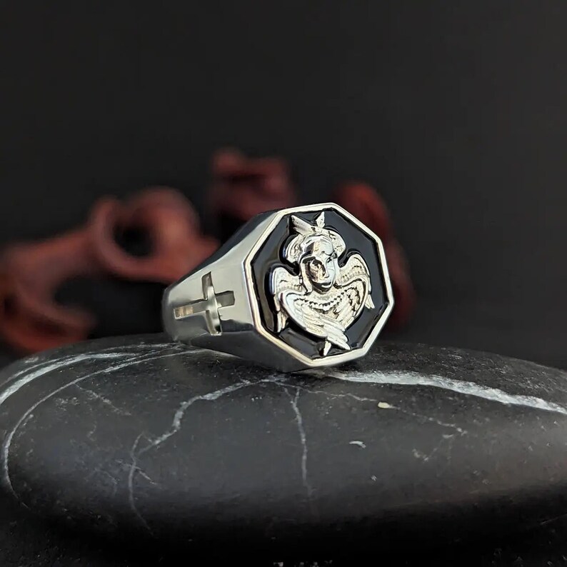 K&ouml;nnte beinhalten: Ein silberner Ring mit einem schwarzen achteckigen Gesicht, das ein detailliertes Engeldesign aufweist. Der Ring hat ein Kreuz an der Seite und ist vor einem dunklen Hintergrund platziert.