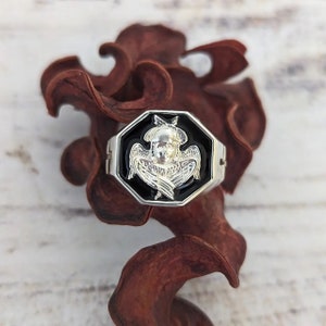 K&ouml;nnte beinhalten: Silberfarbener Ring mit achteckiger schwarzer Fassung, die ein detailliertes Engeldesign zeigt. Der Engel hat ausgebreitete Fl&uuml;gel und einen Heiligenschein. Der Ring ist vor einem verschwommenen Hintergrund aus getrocknetem, r&ouml;tlich-braunem Pflanzenmaterial platziert.