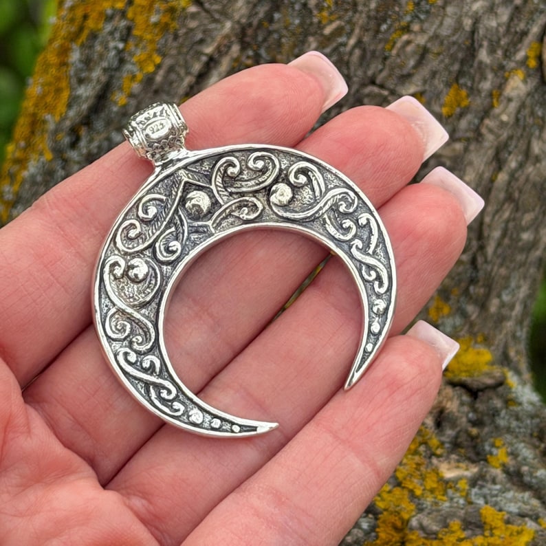 May include: A silver crescent moon pendant with intricate scrollwork and a small loop at the top. The pendant is held in a hand, showcasing the detailed design and texture of the metal. The pendant is marked with "925".