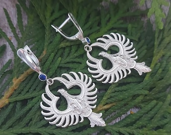 May include: A pair of silver earrings with a detailed design of a bird with spread wings. Each earring features a blue gemstone.