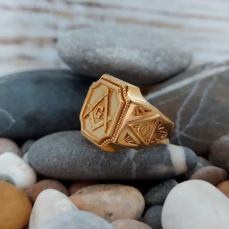 K&ouml;nnte beinhalten: Ein goldener Freimaurer-Ring mit einem Quadrat- und Zirkel-Design und einem allsehenden Auge auf dem Band.