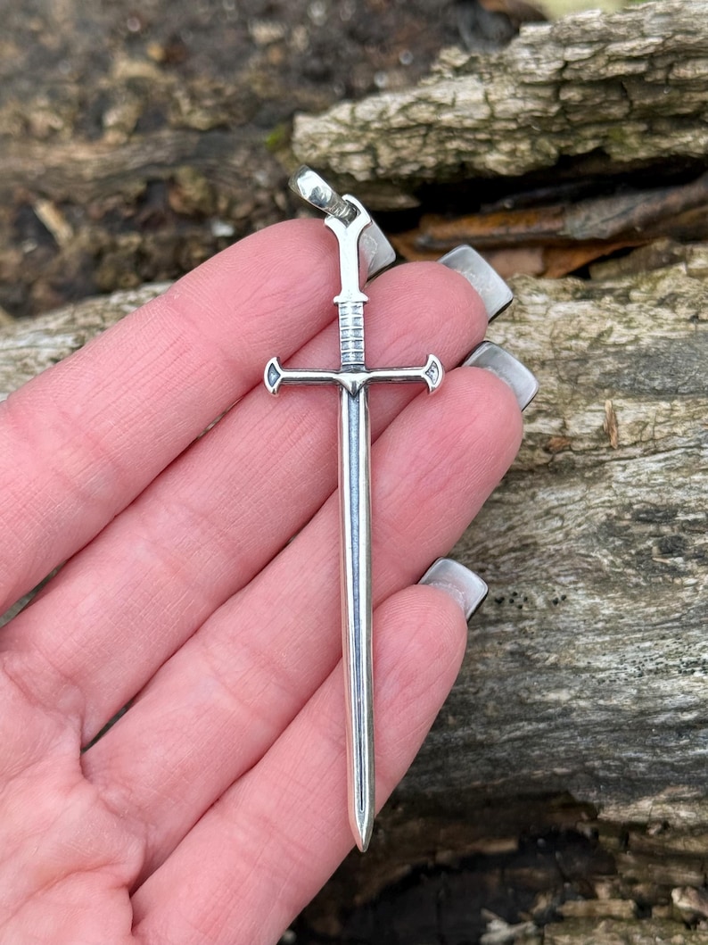 May include: A silver sword pendant with a cross-shaped hilt and a pointed tip.