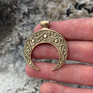 May include: A gold-colored crescent moon pendant with intricate scrollwork and a small loop at the top for a chain. The pendant has a detailed, antique design, and is likely made of brass or a similar metal.