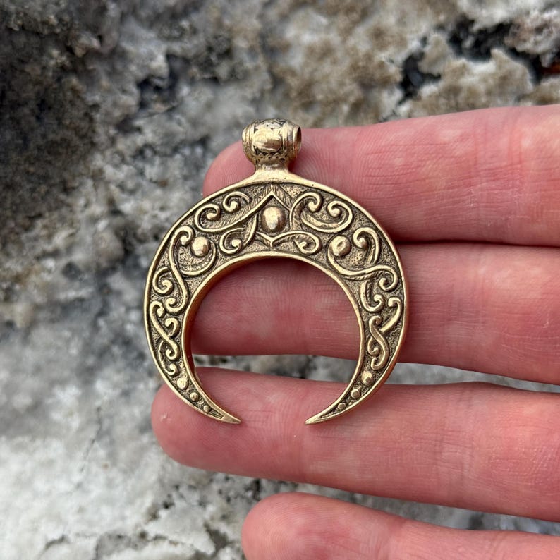 May include: A gold-colored crescent moon pendant with intricate scrollwork and a small loop at the top for a chain. The pendant has a detailed, antique design, and is likely made of brass or a similar metal.
