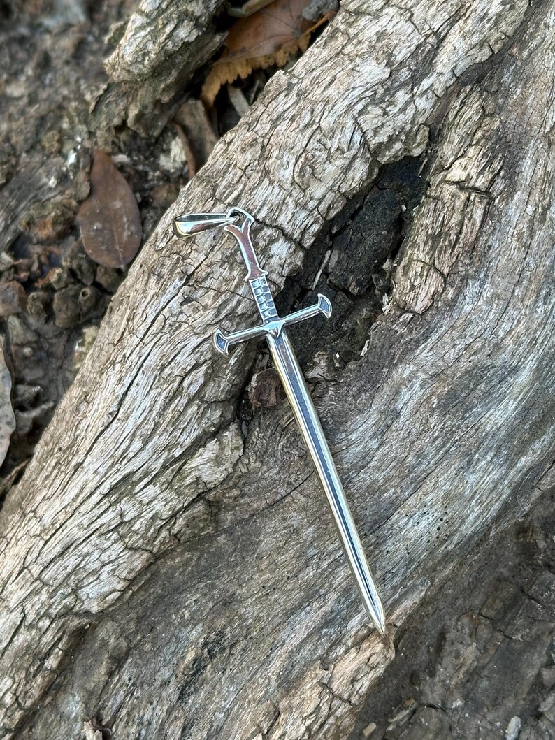 May include: A silver sword pendant with a detailed hilt and a sharp point. The pendant is lying on a piece of weathered wood.
