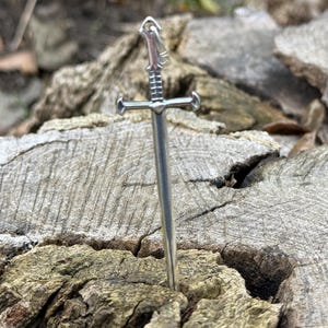 May include: A silver sword pendant with a detailed hilt and a sharp point. The pendant is resting on a piece of weathered wood.