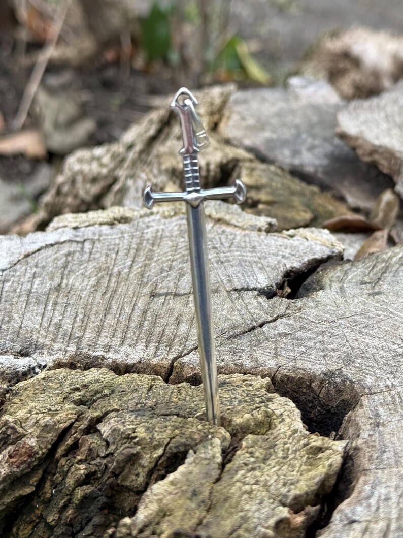 May include: A silver sword pendant with a detailed hilt and a sharp point. The pendant is resting on a piece of weathered wood.