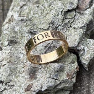 Könnte beinhalten: Ein goldfarbener Ring mit der Inschrift "IS FORTU" um das Band graviert. Der Ring befindet sich vor einem strukturierten, grau-braunen Hintergrund, möglicherweise Holz oder Stein. Die Oberfläche des Rings reflektiert Licht.