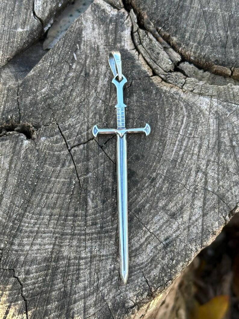 May include: A silver sword pendant with a cross-shaped hilt and a pointed tip. The pendant is on a wooden surface.