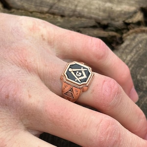 K&ouml;nnte beinhalten: Ein goldener Ring mit einem schwarzen Emaille-Symbol der Freimaurer, das einen Zirkel und ein Geodreieck sowie ein allsehendes Auge zeigt.