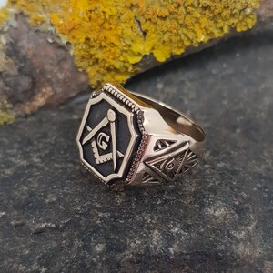 K&ouml;nnte beinhalten: Ein goldener Freimaurer-Ring mit einem schwarzen Emaille-Quadrat und Kompass-Design. Der Ring hat ein detailliertes Design mit einem allsehenden Auge und anderen Freimaurer-Symbolen.