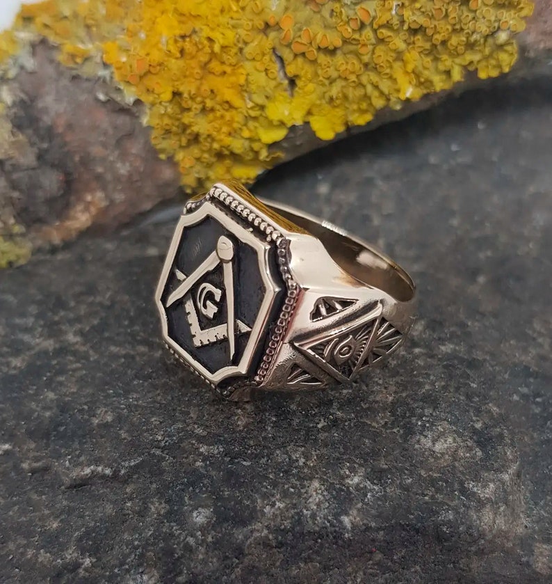K&ouml;nnte beinhalten: Ein goldener Freimaurer-Ring mit einem schwarzen Emaille-Quadrat und Kompass-Design. Der Ring hat ein detailliertes Design mit einem allsehenden Auge und anderen Freimaurer-Symbolen.