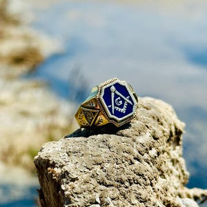 K&ouml;nnte beinhalten: Ein goldener Ring mit einem blauen emaillierten Freimaurer-Symbol. Das Symbol zeigt einen Zirkel und ein Geodreieck mit einem 'G' in der Mitte.