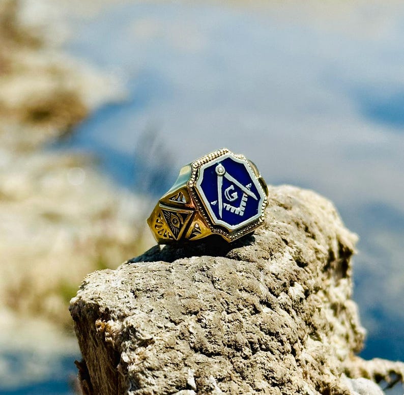 K&ouml;nnte beinhalten: Ein goldener Ring mit einem blauen emaillierten Freimaurer-Symbol. Das Symbol zeigt einen Zirkel und ein Geodreieck mit einem 'G' in der Mitte.