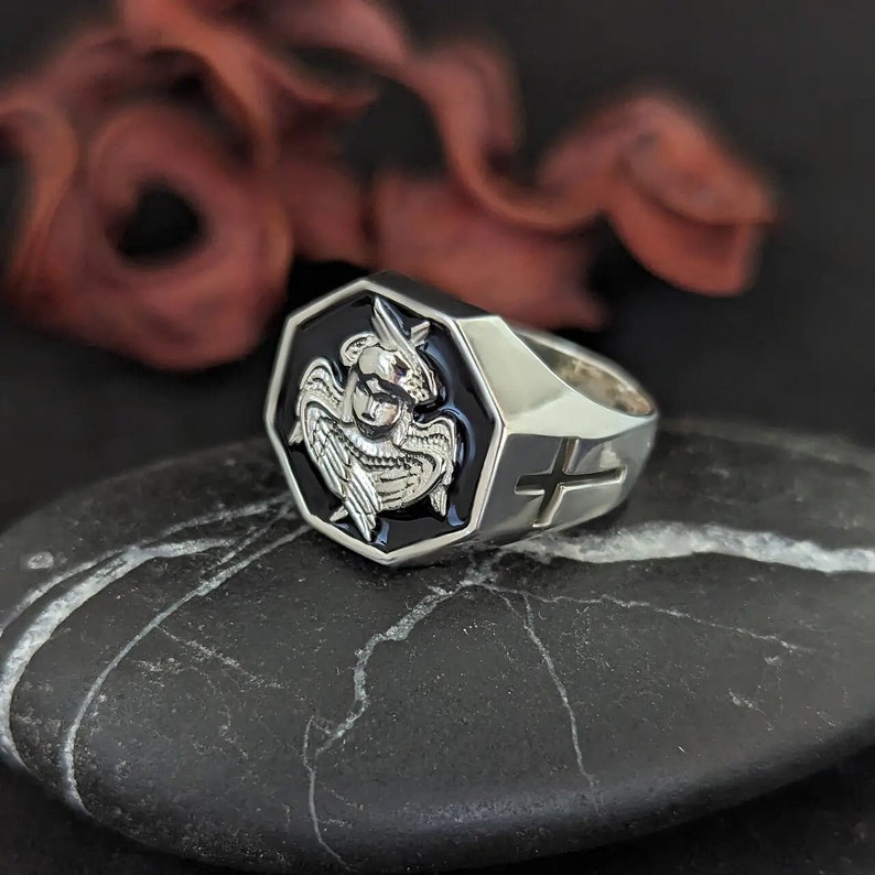 K&ouml;nnte beinhalten: Ein silberner Ring mit einer schwarzen sechseckigen Fassung, die ein detailliertes Relief einer gefl&uuml;gelten Figur zeigt. Der Ring hat eine polierte Oberfl&auml;che und ein Kreuzdesign an der Seite. Der Ring wird auf einer dunklen, strukturierten Oberfl&auml;che pr&auml;sentiert.