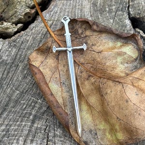 May include: A silver sword pendant with a detailed design, resting on a brown leaf. The pendant is a popular choice for jewelry and is often used as a symbol of strength and courage.
