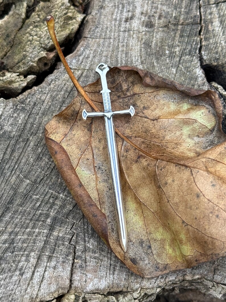 May include: A silver sword pendant with a detailed design, resting on a brown leaf. The pendant is a popular choice for jewelry and is often used as a symbol of strength and courage.