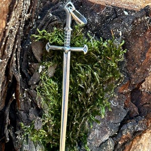 May include: A silver sword pendant with a cross guard and a loop at the top for attaching to a chain. The pendant is resting on a bed of green moss.