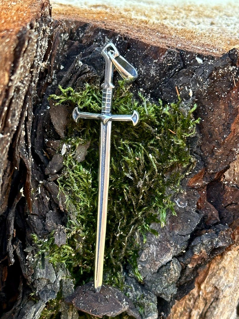 May include: A silver sword pendant with a cross guard and a loop at the top for attaching to a chain. The pendant is resting on a bed of green moss.