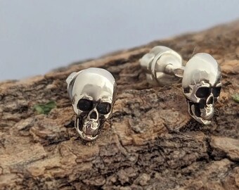Può includere: Orecchini a lobo a forma di teschio in argento. Gli orecchini sono piccoli, dettagliati e presentano un design a teschio. I teschi hanno orbite scure e sono posti su uno sfondo di legno naturale.