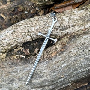 May include: A silver sword pendant with a detailed design. The pendant is hanging from a chain and is resting on a piece of weathered wood.