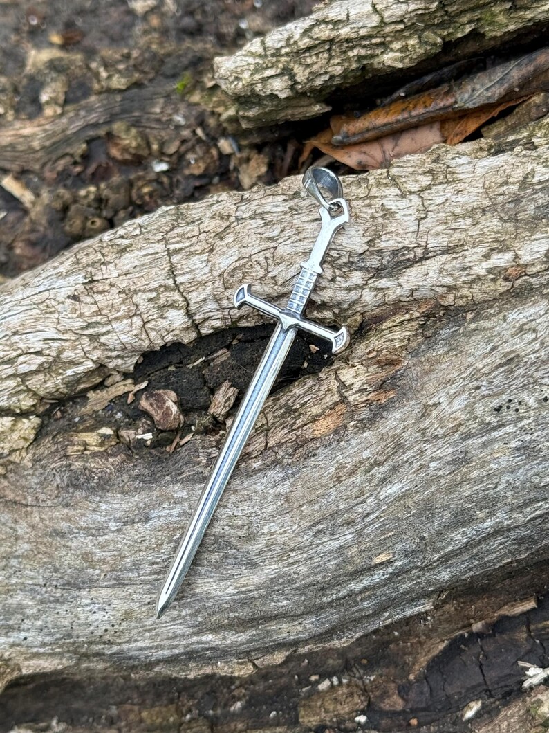 May include: A silver sword pendant with a detailed design. The pendant is hanging from a chain and is resting on a piece of weathered wood.