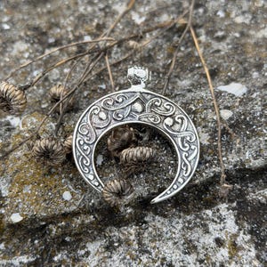 May include: A silver-colored crescent moon pendant with intricate scrollwork and a small loop for a chain. The pendant is displayed on a textured gray stone surface, with dried plant stems and seed heads in the background.