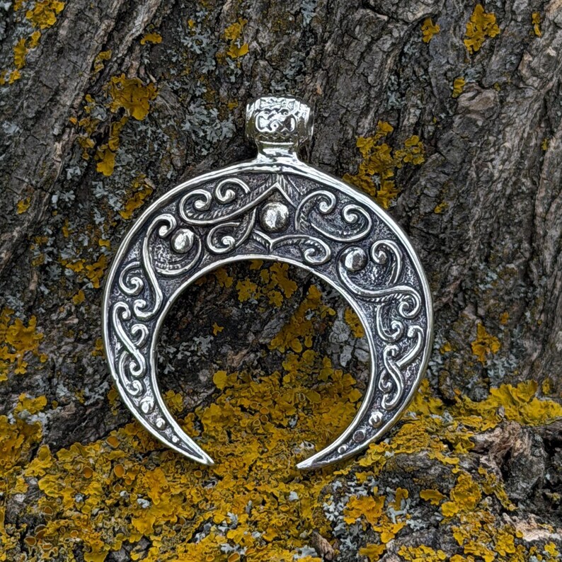 May include: Silver-toned crescent moon pendant with intricate scrollwork and small beaded accents. The pendant has a crown-like detail at the top and is displayed against a textured, natural background.